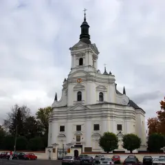 Biała świątynia z wieżą, gdzie czczona jest Matka Boska Kodeńska. Otoczona drzewami i parkingiem.