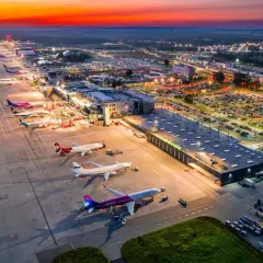 Zachód słońca nad lotniskiem Katowice Airport. Samoloty czekają na pasie startowym, a parkingi są pełne samochodów.