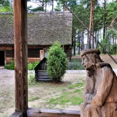 Drewniana rzeźba postaci, tradycyjna chata z mchem na dachu i zielone drzewa tworzą klimatyczne **kadzidło atrakcje** w skansenie.