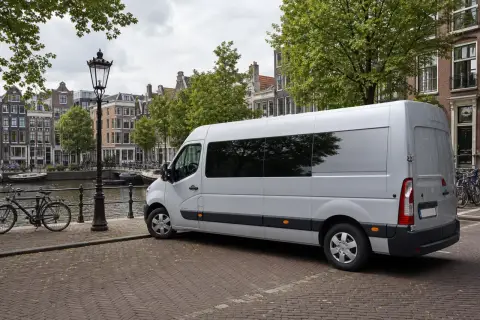 Biały bus czeka na załadunek, gotowy do transportu rzeczy z adresu pod adres. W tle kanał i kamienice Amsterdamu.