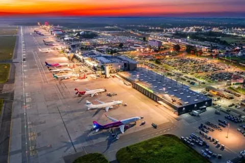 Zachód słońca nad lotniskiem Katowice Airport. Samoloty czekają na pasie startowym, a parkingi są pełne samochodów.