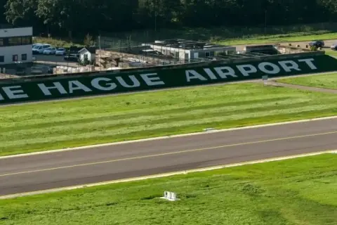 The Hague Airport z wielkimi białymi literami na zielonym tle. Pas startowy i budynki lotniska.