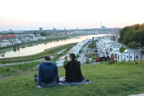 Dwa osoby siedzą na trawie, podziwiając panoramę nowoczesnego osiedla nad wodą w Dortmundzie.
