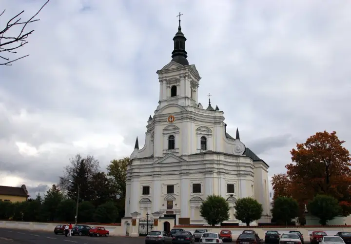 Biała świątynia z wieżą, gdzie czczona jest Matka Boska Kodeńska. Otoczona drzewami i parkingiem.