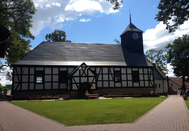 Kościół z wieżą zegarową, otoczony zielenią i ławkami. W tle niebo z chmurami. Miejsce pielgrzymek do Matki Boskiej Sianowskiej.