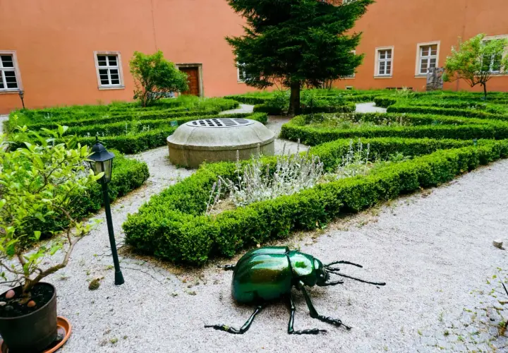 Ogród klasztorny z rzeźbą zielonego chrząszcza, studnią i żywopłotami.