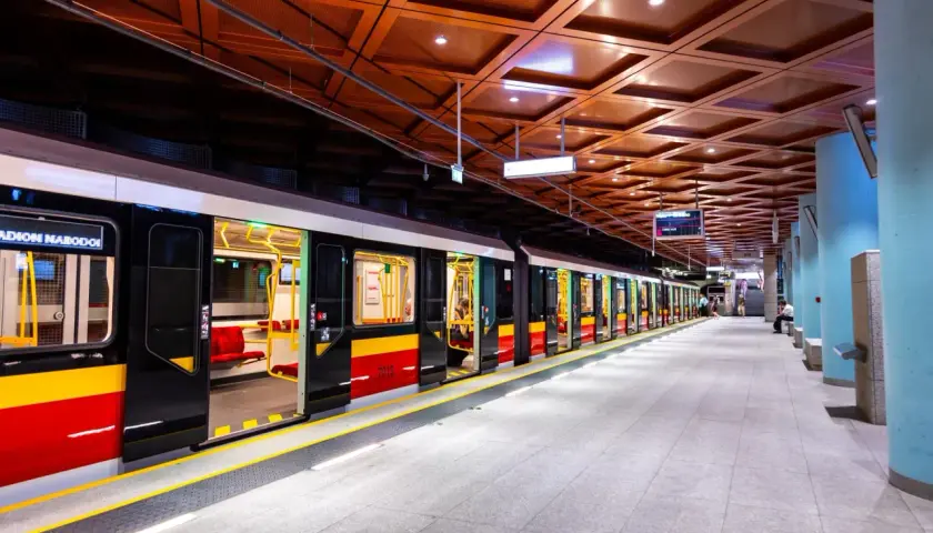 Nowoczesny pociąg na jednej z warszawskich stacji metra M2. Jasne wnętrze wagonów i charakterystyczne czerwono-żółte pasy na zewnątrz.