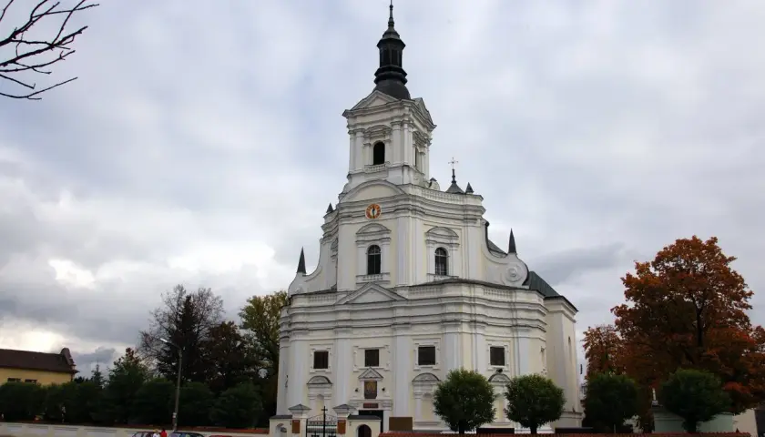 Biała świątynia z wieżą, gdzie czczona jest Matka Boska Kodeńska. Otoczona drzewami i parkingiem.