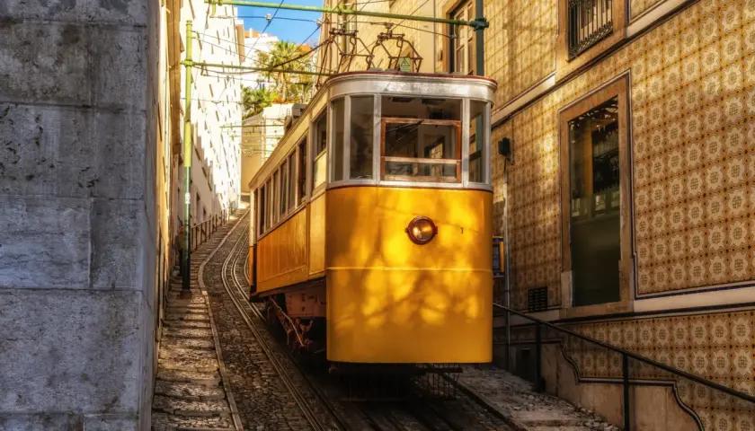 Żółty tramwaj wspina się po stromych torach w wąskiej uliczce Lizbony. W tle widać kamienne budynki pokryte tradycyjnymi płytkami azulejo.