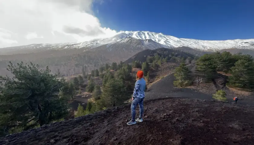 Kobieta w pomarańczowej czapce patrzy na ośnieżony wulkan Etna, otoczony lasem i czarną ziemią.