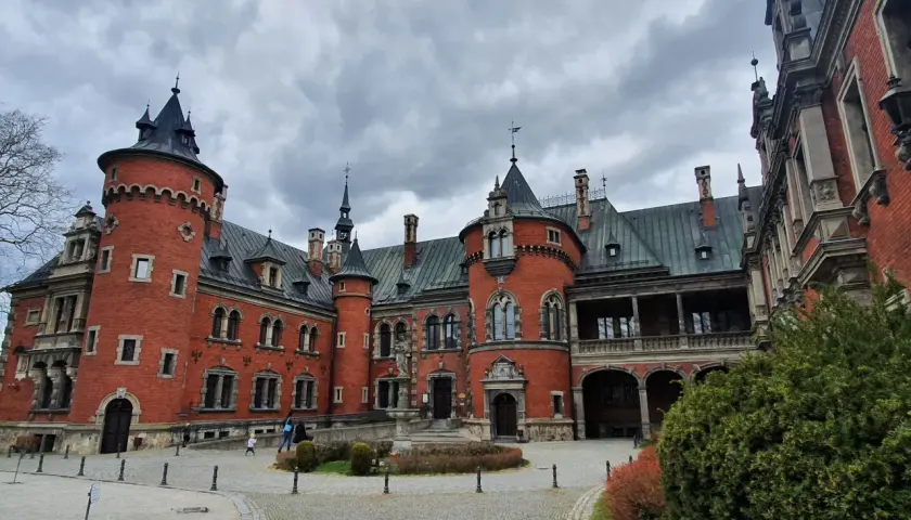 Pławniowice atrakcje: okazały zamek z czerwonej cegły z wieżami i arkadowym gankiem, otoczony brukowanym dziedzińcem i zielenią.