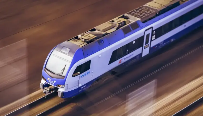 Białystok elk pędzi po torach. Nowoczesny pociąg Stadler PKP Intercity z niebiesko-białymi pasami mknie przez krajobraz.