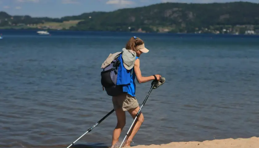 Osoba z plecakiem i kijkami do nordic walking nad morzem, wchodząca do wody.