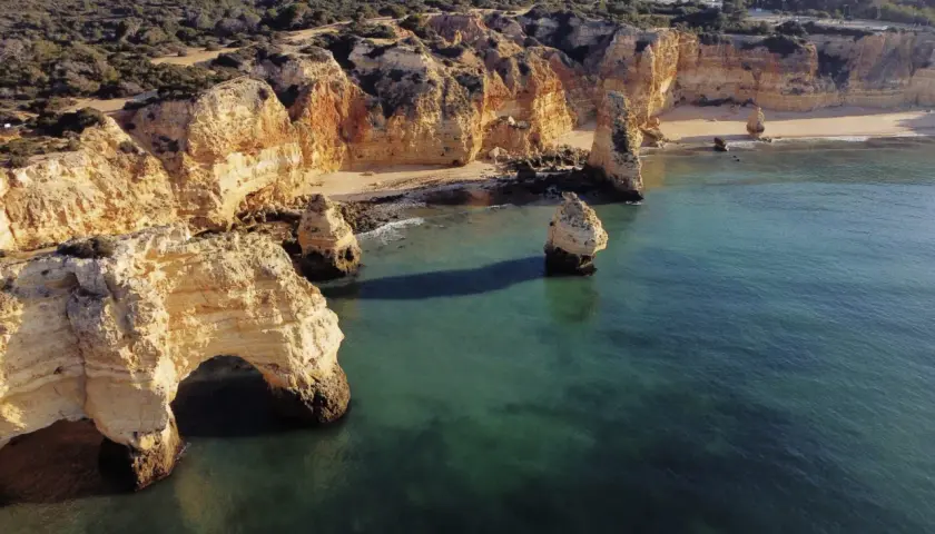 Skaliste klify Algarve, z łukiem skalnym i samotnymi formacjami, wpadają do turkusowego morza. Widok przypomina o urokach wybrzeża, jakbyśmy byli w pobliżu Faro Lizbona.