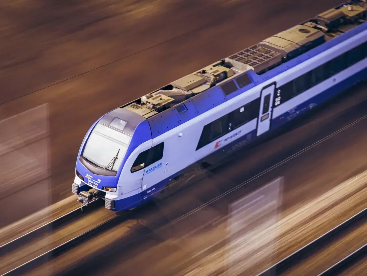 Białystok elk pędzi po torach. Nowoczesny pociąg Stadler PKP Intercity z niebiesko-białymi pasami mknie przez krajobraz.