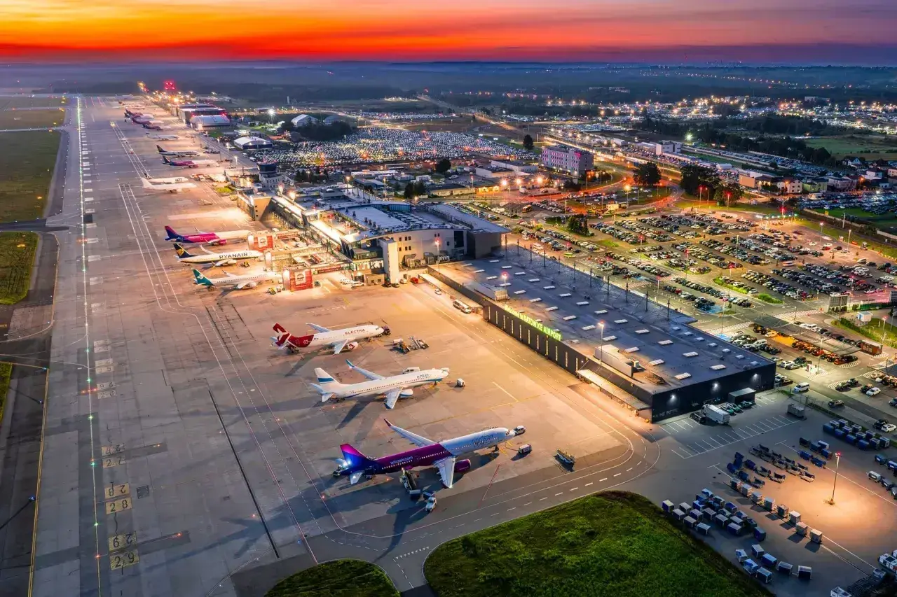 Zachód słońca nad lotniskiem Katowice Airport. Samoloty czekają na pasie startowym, a parkingi są pełne samochodów.