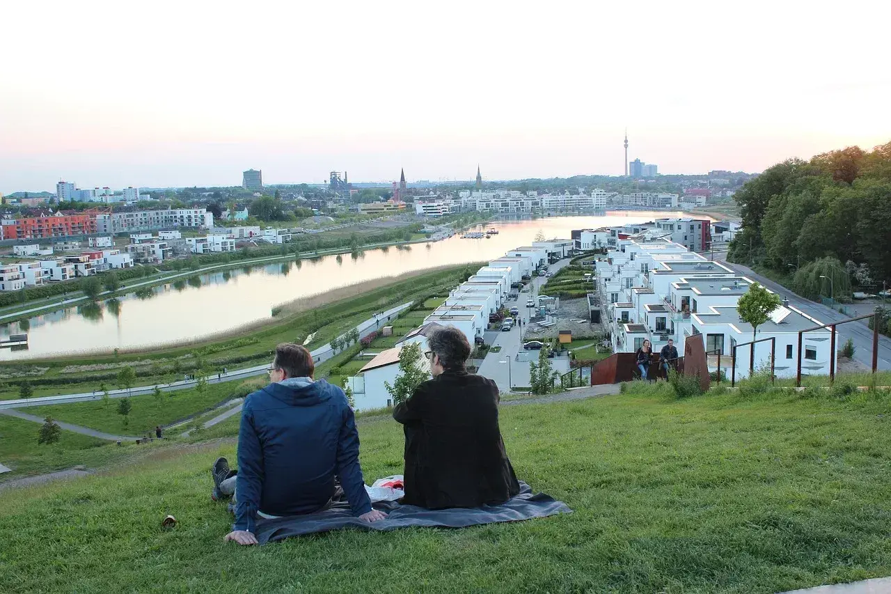 Dwa osoby siedzą na trawie, podziwiając panoramę nowoczesnego osiedla nad wodą w Dortmundzie.