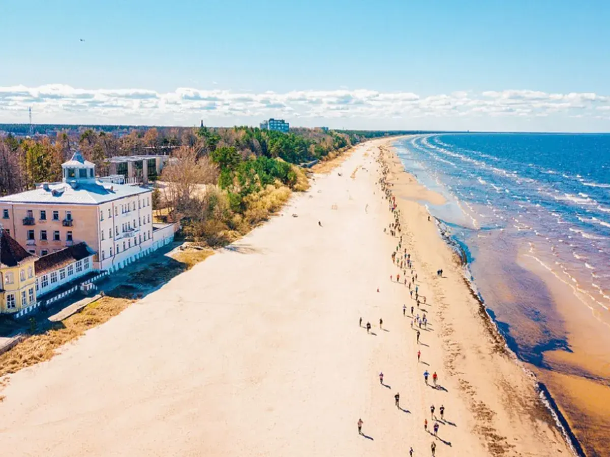 Szeroka, piaszczysta plaża na Łotwie, z budynkami przy brzegu i ludźmi spacerującymi wzdłuż morza.