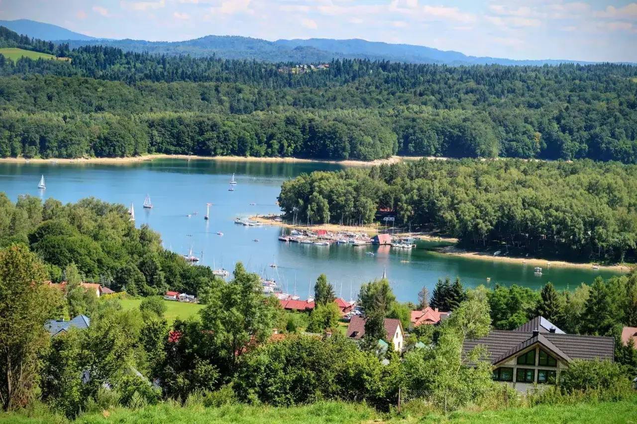 Malowniczy widok na jezioro z żaglówkami i lasami. Idealne miejsce na wakacje, łatwo tu dojechać nad Solinę samochodem.