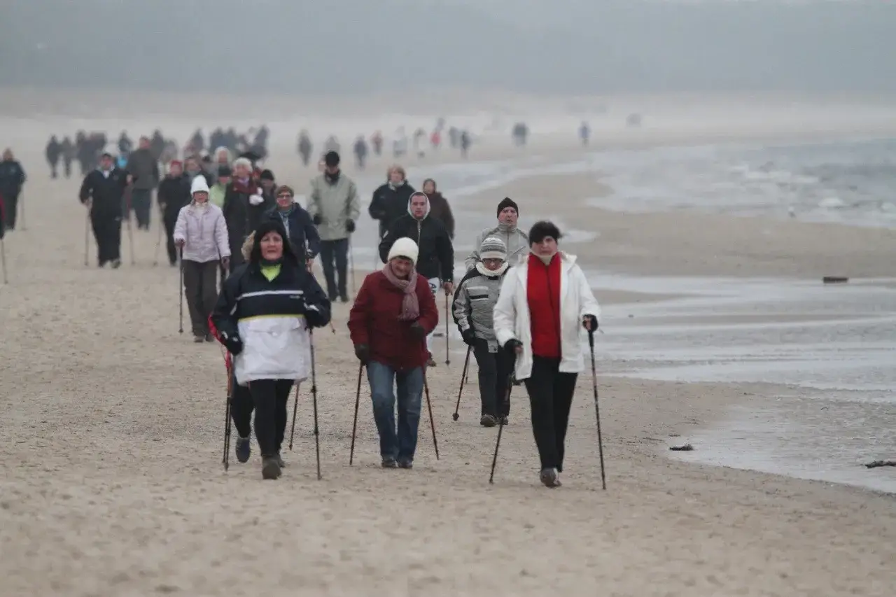Grupa osób uprawia nordic walking nad morzem. Spacerują po piaszczystej plaży w chłodny, mglisty dzień.